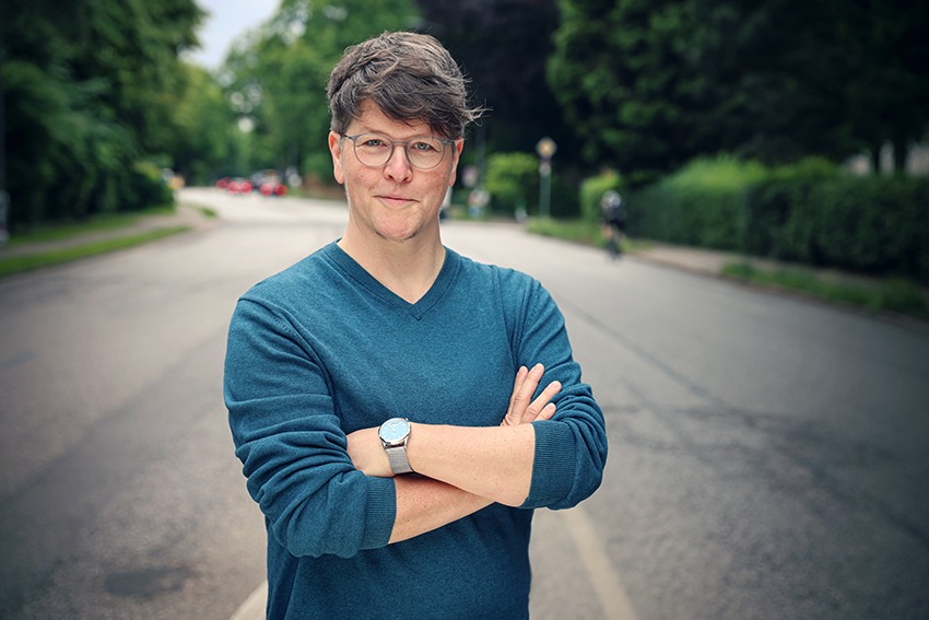 Jona trägt einen petrolfarbenen Pullover und hat kurze dunkle Haare. Jona blickt mit verschränkten Armen leicht schmunzelnd in die Kamera.<br />
© Carina Häusler