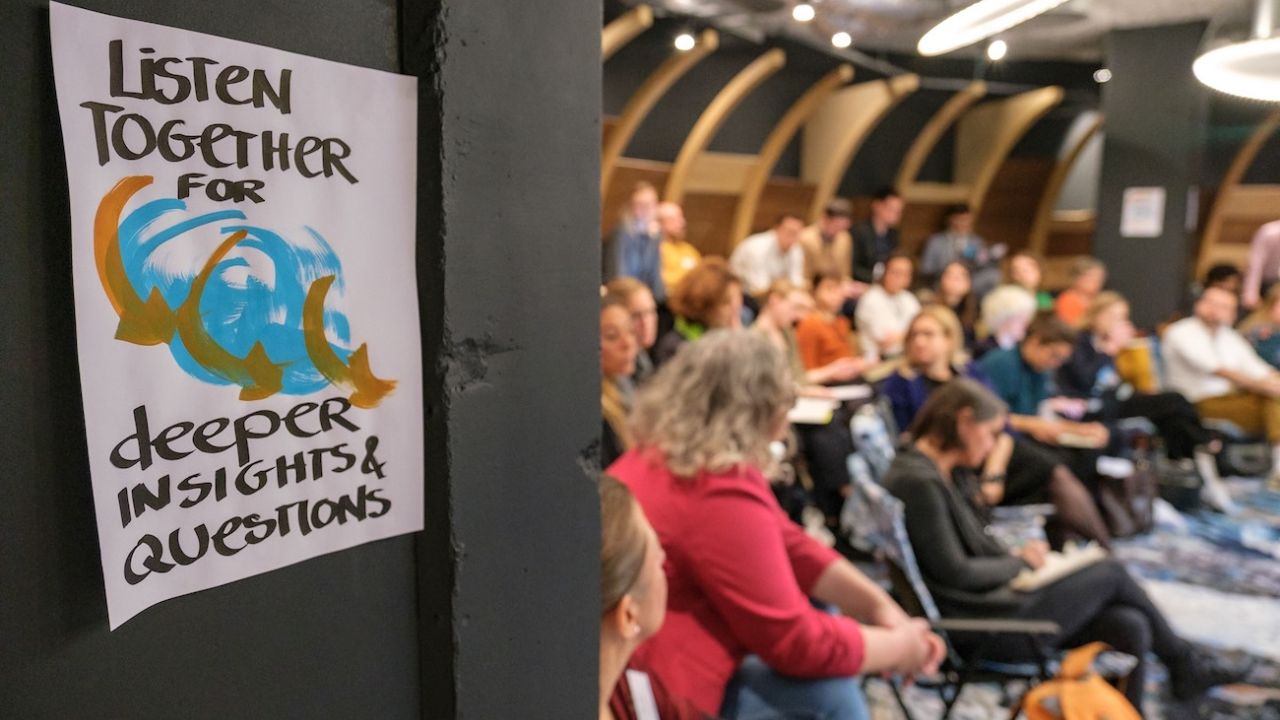Ein hangeschriebenes Plakat mit dem Text "listen together for deeper insights & questions" hängt am Eingang zu einem Raum, in dem sich viele Menschen befinden
